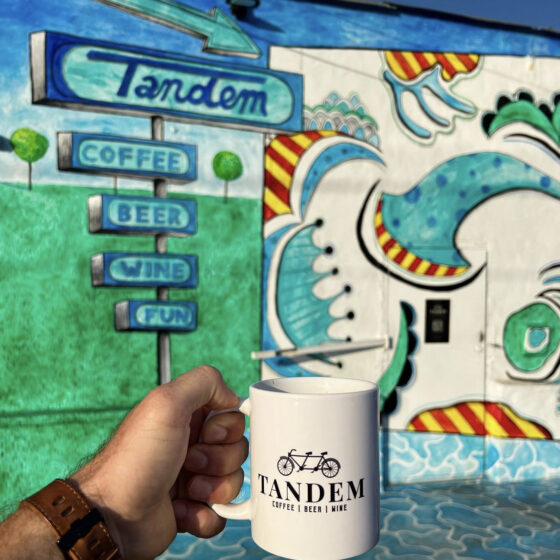 A coffee mug with the tandem logo appears in front of colorful coffee shop in San Antonio.