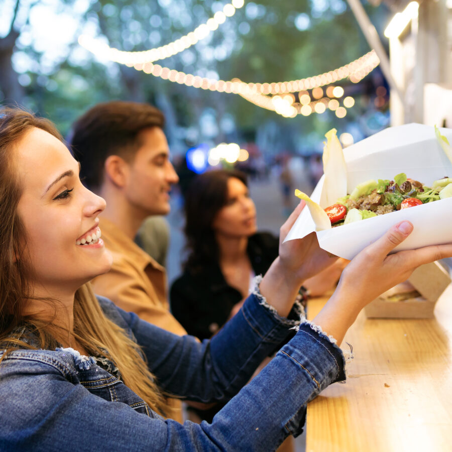 Group of attractive young friends choosing and buying different types of fast food at food trucks in the town center.