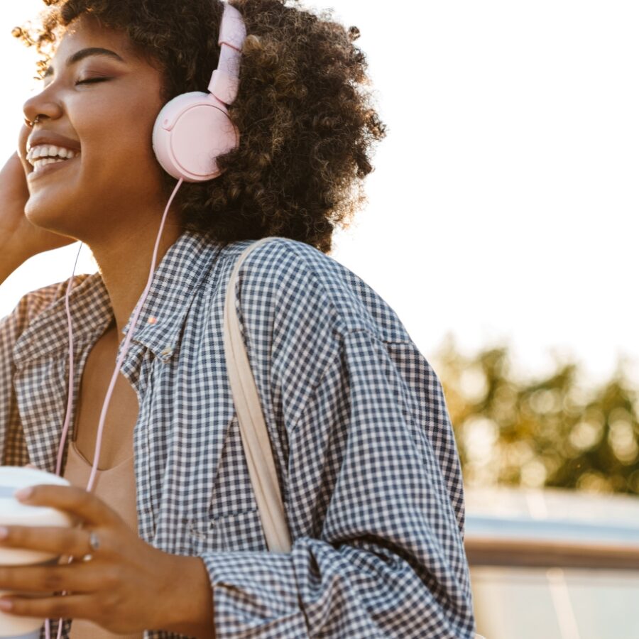 Curly haired young woman listens to music on pink headphones while walking through the town center and drinking a to-go coffee.