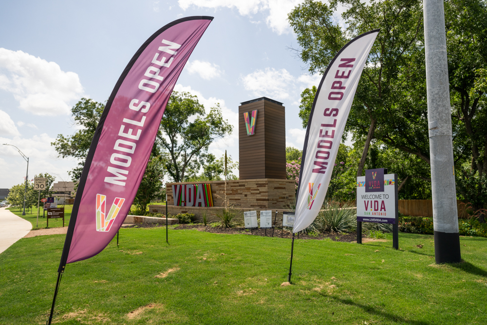 "Model Homes Now Open" flags wave in front of colorful VIDA San Antonio signage at the community entrance.