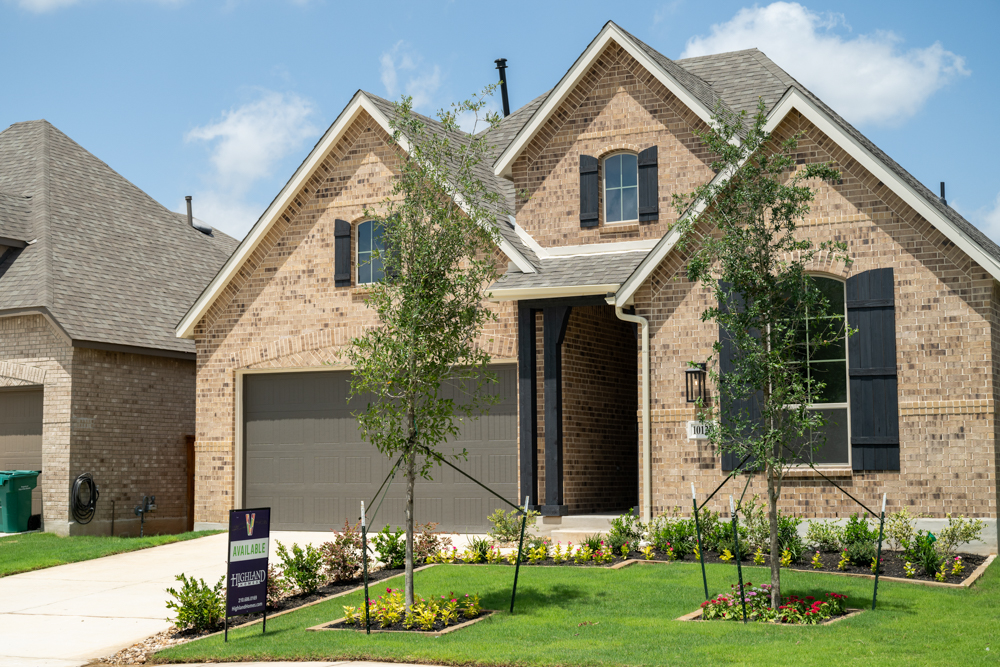 A newly constructed Highland Home for sale with brick walls, a lush front yard, young trees, and flowers