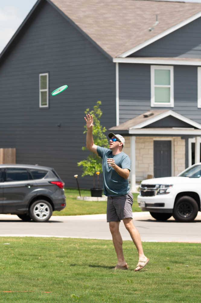 A father wearing a hat and sunglasses plays frisbee in the neighborhood park.