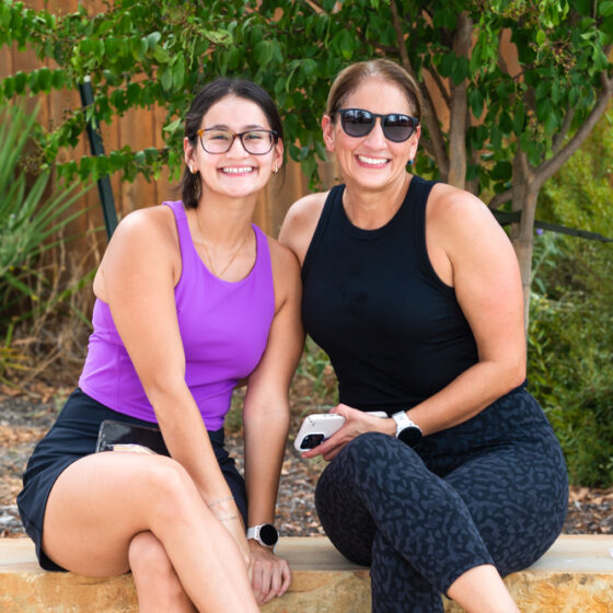 A mother and daughter in athletic wear sit against a green backdrop and smile