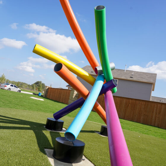 A public artwork with colorful tubes stands on a black pedestal on a green lawn.