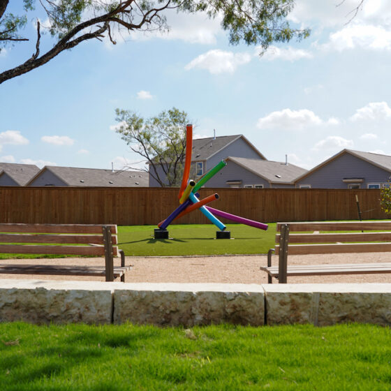 Two wooden park benches in front of a vibrant piece of public art in San Antonio. It stands in a VIDA San Antonio park with a green lawn and a tall tree, houses in the background.