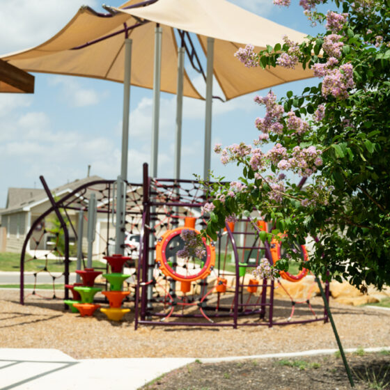 A park scene within the VIDA San Antonio community showcases a playground with climbing structures under shade sails. A pergola offers shade on the concrete walkway, surrounded by crepe myrtle trees and grassy areas. Charming houses are visible in the background beneath a cloudy blue sky.