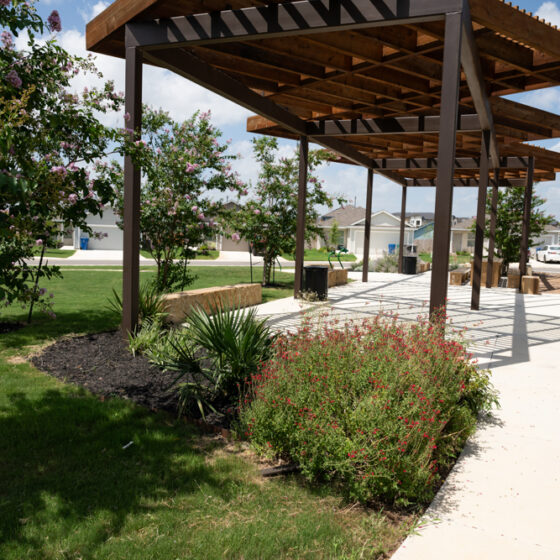 A park scene within the VIDA San Antonio community showcases a playground with climbing structures under shade sails. A pergola offers shade on the concrete walkway, surrounded by trees and green grassy areas. Charming houses are visible in the background beneath a clear blue sky.