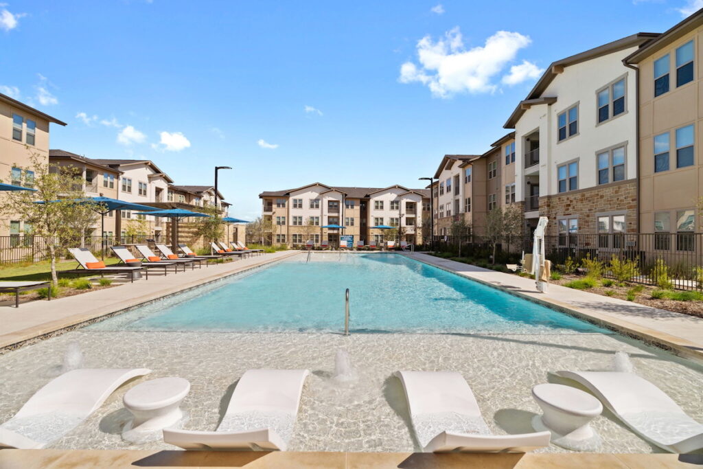 A crystal blue pool with white pool chaises in the middle of a modern apartment community on a sunny day