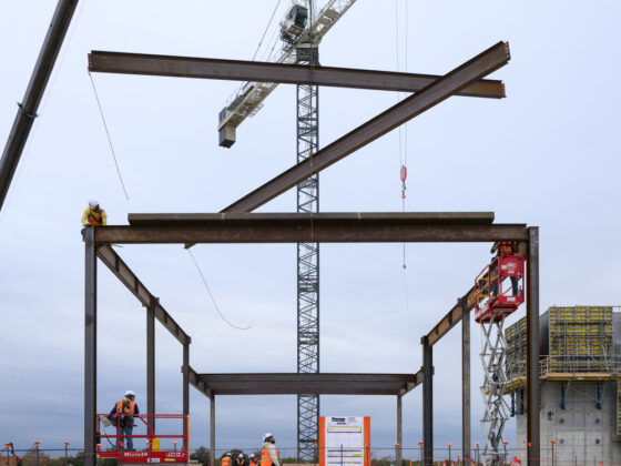 Several beams of steel hang in the sky as construction workers use a crane to lay beams onto metal frame