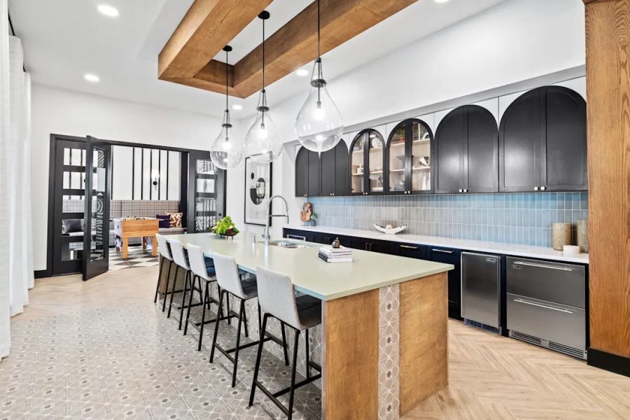 Modern kitchen with a long island, bar stools, and pendant lights. It features dark arch-shaped cabinets, a light countertop, light wood accents, and a herringbone floor. A foosball table is visible in the adjoining room.